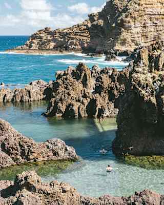 Three swimmers enjoy the pristine sea pools created by rocky outcrops along the coastline at Porto Moniz, seen from above.