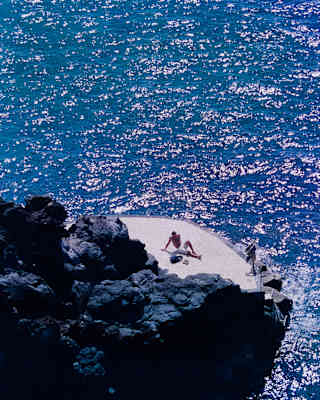 A man lies back on a swimming platform at the base of volcanic rocks, surrounded by glittering azure sea, seen from above.