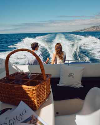 A man and woman sit at the back of a boat on the sea, facing the wake. A wicker picnic basket and a book are on a table in the foreground under a sunny sky.