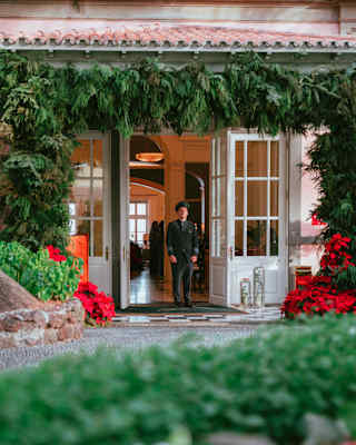 A member of staff in dark uniform stands at the entrance under a festive decoration garland of fir, seen from gardens.