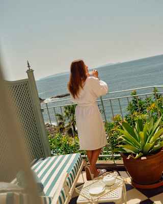 A woman in a robe drinks tea with elbows resting on the railing of a private terrace overlooking the sea, seen from behind.