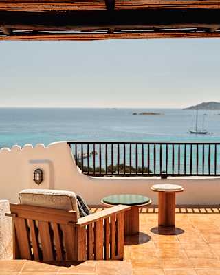 A cushioned, wood-framed terrace chair with two small side tables faces the sublime Costa Smeralda sea, seen from behind.