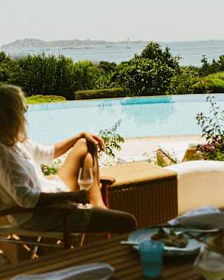 A guest in cool white sits in shade with a glass of wine, seen in soft-focus, gazing over a villa pool to the sea beyond.