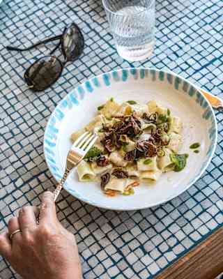 A diner holds a fork above a bowl of mezzi paccheri alla Nerano at a mosaic-tiled table with a water glass and sunglasses.