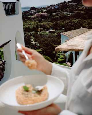 In soft-focus foreground a woman holds a bowl of risotto with tomato water, with a backdrop of Romazzino's verdant grounds.