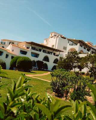 Angled image of Romazzino's white exterior with arch and rhombus windows and roof terraces, seen from lush green gardens.