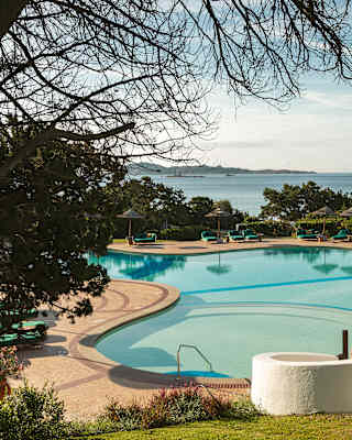 Looking down from green sloping gardens to the tropical pool below, with views past the sun-loungers and parasols to the sea.