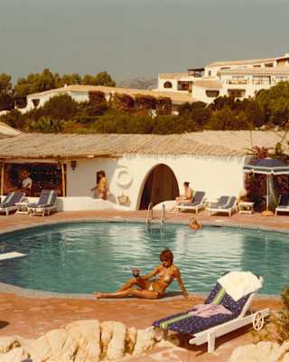 Historic honeyed Kodachrome photo of guests lounging with drinks by the sun-drenched resort pool, with a thatched bar area.