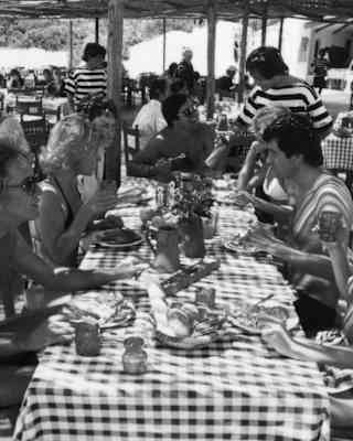 Historic black and white image of the busy terrace, as guests lunch at sun-dappled tables, helped by waiters in striped tops.