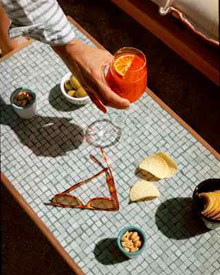 A hand places a Negroni cocktail between bowls of crisps, nuts, olives and a pair of sunglasses on a grey mosaic table top.