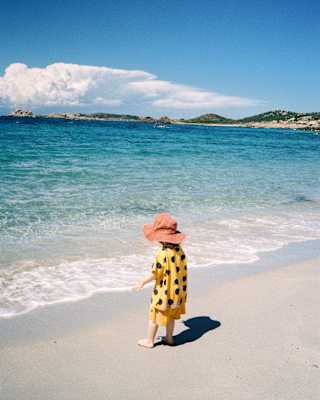 A small child in a yellow and black spotty top and red sunhat approaches the white foam shallows of the bright turquoise sea.