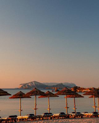 Sun glows on thatched parasols and flat sun-loungers lining the waterfront as island contours rise into a soft sunset sky.