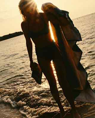 A woman, with a magazine in one hand and towel in the other, stands in white-water shallows, silhouetted against golden sun.