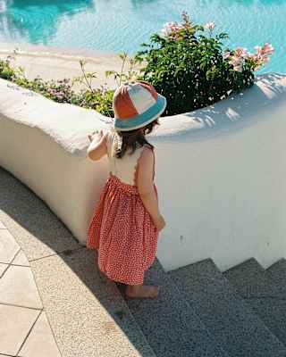 A small girl with a red and white dress and matching striped hat with turquoise brim stands on steps overlooking the pool.