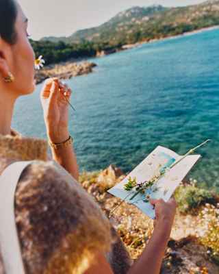 A woman holds a hiking leaflet covered in stems in one hand, and with the other sniffs a daisy-like flower, seen side-on.