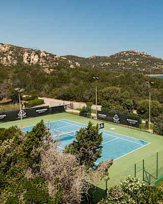 A rocky headland and flashes of bright azure ocean are seen behind a coastal forest, in a view from above the tennis court.