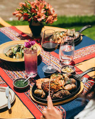 A table set outdoors with colourful patterned cloth, featuring grilled chicken, vegetables, sauces, glasses of wine and water, a pink drink, and a vase of flowers. A person is holding a fork over their plate.