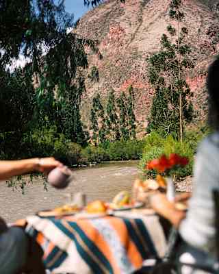 In soft-focus foreground a couple enjoy breakfast overlooking the river, which flows towards a steep, red-brown hillside.
