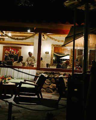 A waiter talks with a couple at a table in El Huerto, illuminated by restaurant lights, viewed through a window from outside.