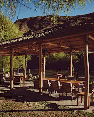 An outdoor wooden pavilion with a tiled roof shelters several tables and chairs on a grassy area, surrounded by trees and mountains under a clear blue sky.
