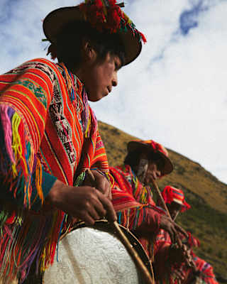 A low-angle shot of a man in a woven Manta as he plays a cumaru wood drum alongside other musicians celebrating Pachamama.