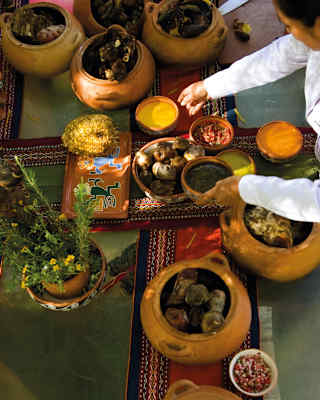 A woman arranges a table with different-sized earthenware bowls of ingredients on a table in dappled light, seen from above.
