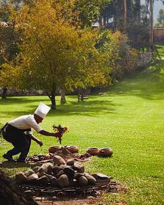 A chef wearing a white hat bends over near a pile of stones and clay pots on a grassy field, surrounded by trees in a sunlit outdoor setting.