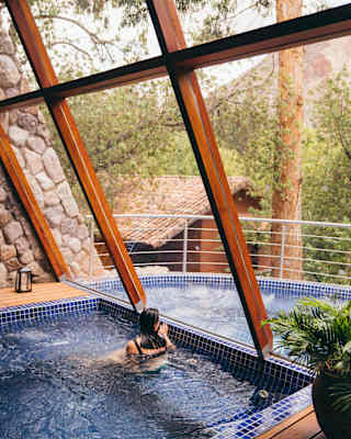 A woman with dark hair relaxes in the bubbling indoor jacuzzi by the window, gazing out on to the steaming outdoor spa pool.