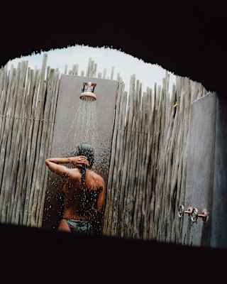 Angled image of a dark-haired woman in an outside shower with a monsoon head and wood fence wall, seen from behind.