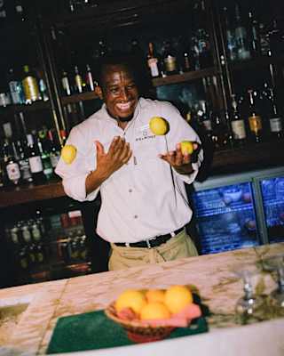 A barman at Eagle Island Lodge laughs as he juggles three lemons behind the marble-top bar, with bottles shelves behind.