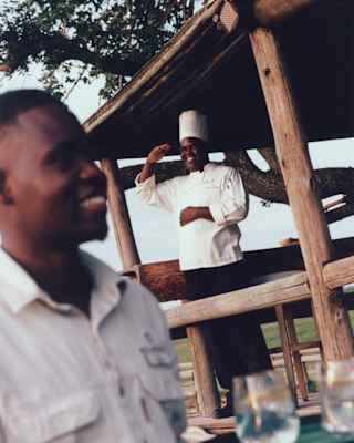 A chef in a toque smiles and salutes from the Fish-Eagle Bar, while a waiter with a tray passes by in soft-focus foreground.