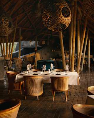 A wicker pendant lamp hangs in the restaurant, complementing wood beams and chairs, seen around a laid white-clothed table.
