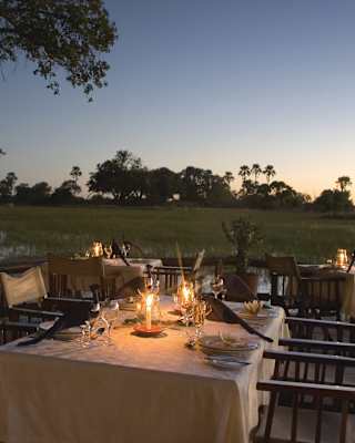 Outdoor dining setup at sunset, with tables and chairs arranged on a patio, candles lit on the tables, overlooking a serene grassy landscape with trees in the distance.