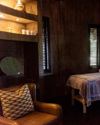 Two treatment beds await guests with rolled towels in a low-lit spa room with four slatted windows and backlit shelves.