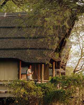 A male guest enjoys a glass of wine on the balcony of his thatched lodge, seen through trees from the exterior.