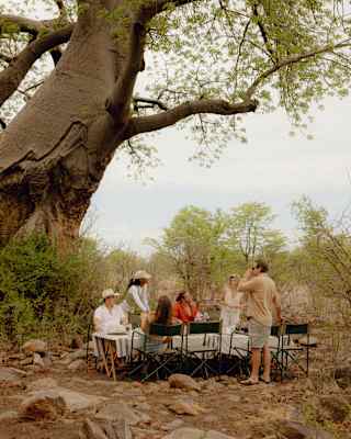 Three men and three women enjoy lunch on fold-out tables and chairs on the stone-studded earth beneath a giant baobab tree.