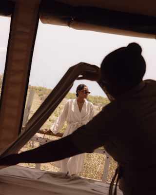 A woman in a white robe and shades leans on the deck rails at the spa as a staff member in the foreground arranges towels.