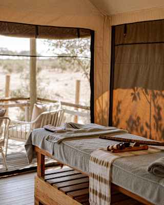 View of a wood-framed spa table with blue towelling top, seen in spa tent with views through netting of an outside deck.