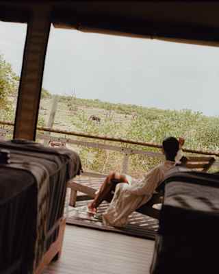 A person in a dressing gown relaxes on a lounger on a covered veranda, overlooking a green, tree-filled landscape through an open balustrade.