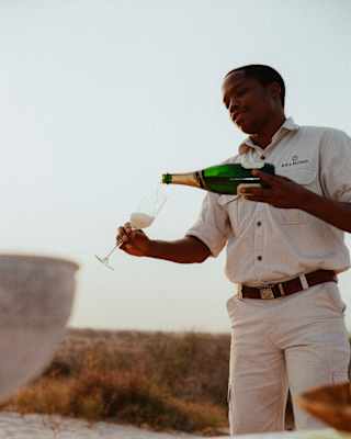 A male member of staff carefully pours Champagne from a bottle into a flute for a sundowner experience, seen at a low-angle.