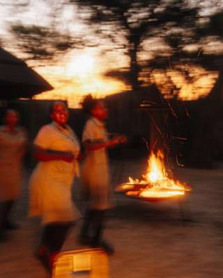 Savute Elephant Lodge staff dance by a fire pit in a clearing, blurred with motion, as the sun sets behind the treeline.