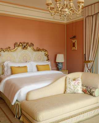 A cream chaise-longue abuts a bed with white linen and gold Rococo frame, flanked by tables against soft coral-pink walls.