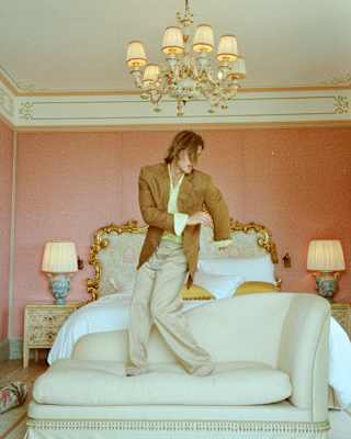 A man in a brown jacket and pale trousers dances on a cream chaise-longue abutting a rococo-framed bed beneath a chandelier.