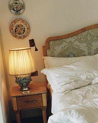 A lamp glows on a table below two decorative wall plates by an unmade bed with white bedding, in a Rivieria Suite detail.