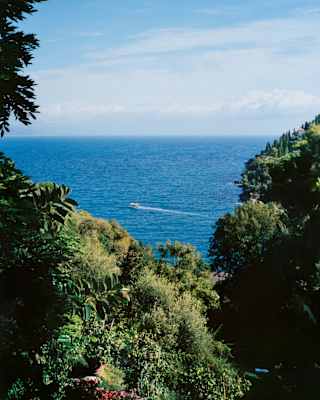 A boat crosses the deep blue sea which meets a lightly clouded horizon in a high-up view between foliage-covered hills.