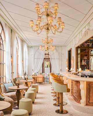 Lamp chandeliers hang above an opulent lounge with luxurious chairs and stools opposite a marble bar with tub-back stools.