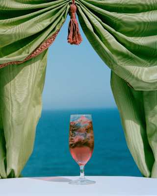 A pink hibiscus-style cocktail with ice sits on a sunny white table overlooking the sea, framed by a green organza curtain.