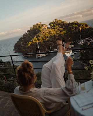 A woman in linens enjoys a glass of wine with her feet up on the terrace railings, gazing at Baia Cannone, seen from behind.
