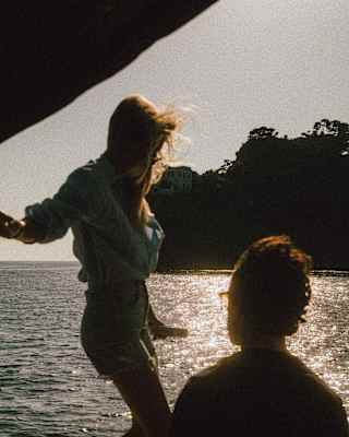 A woman with long hair walks along the side of a boat watched by a friend, both half-silhouetted against the sun-kissed sea.