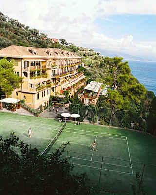 High-angle view of a couple playing tennis on a green court with a backdrop of the hotel nestled in hills overlooking the sea.